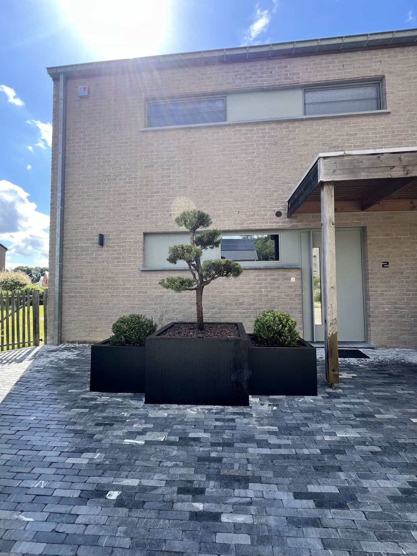 Devanture d'une maison moderne en pavés foncés, avec des bacs noirs et un arbre taillé