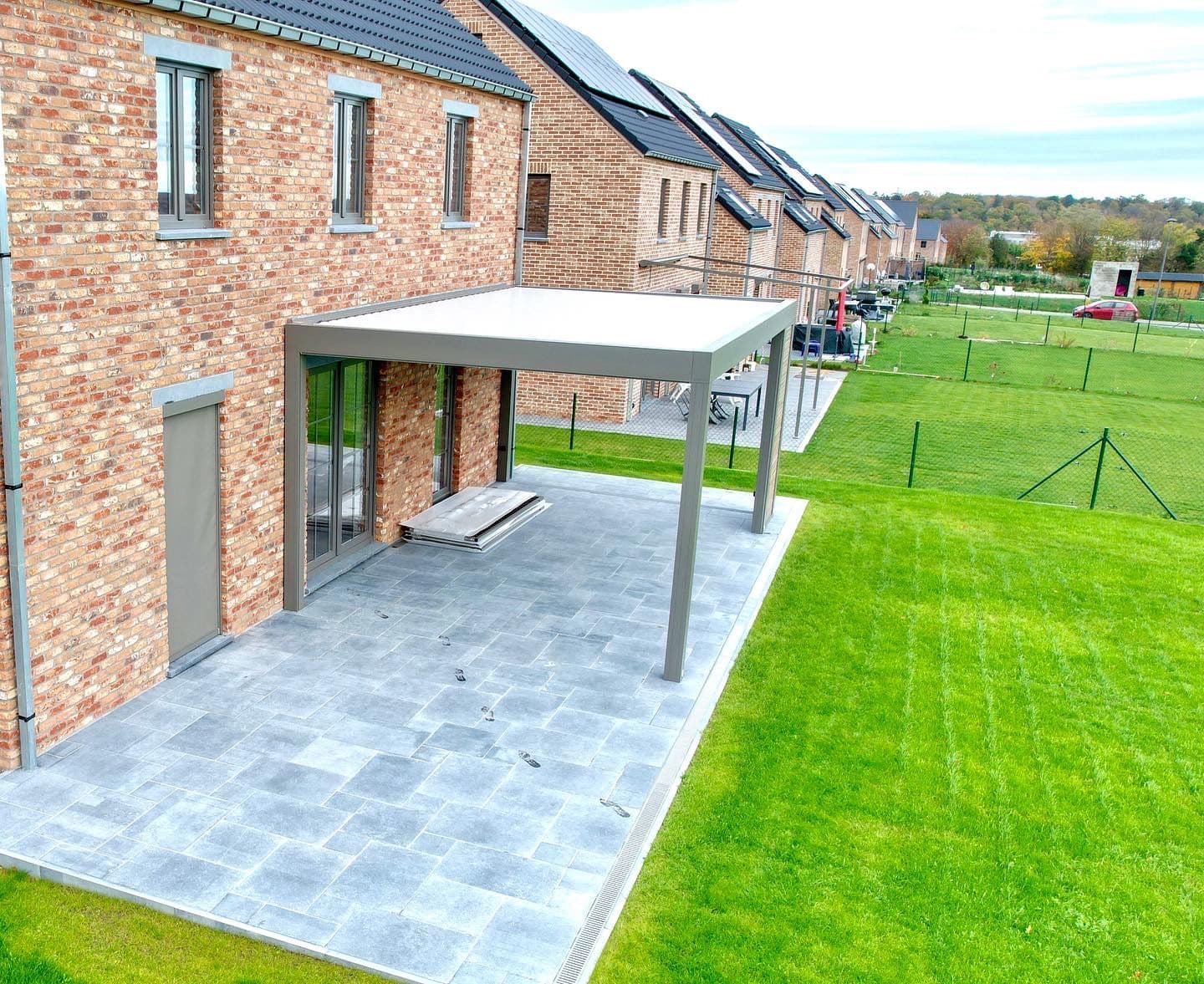 Terrasse en pavés sous une pergola blanche, à l'arrière d'une maison en briques, bordée de pelouse